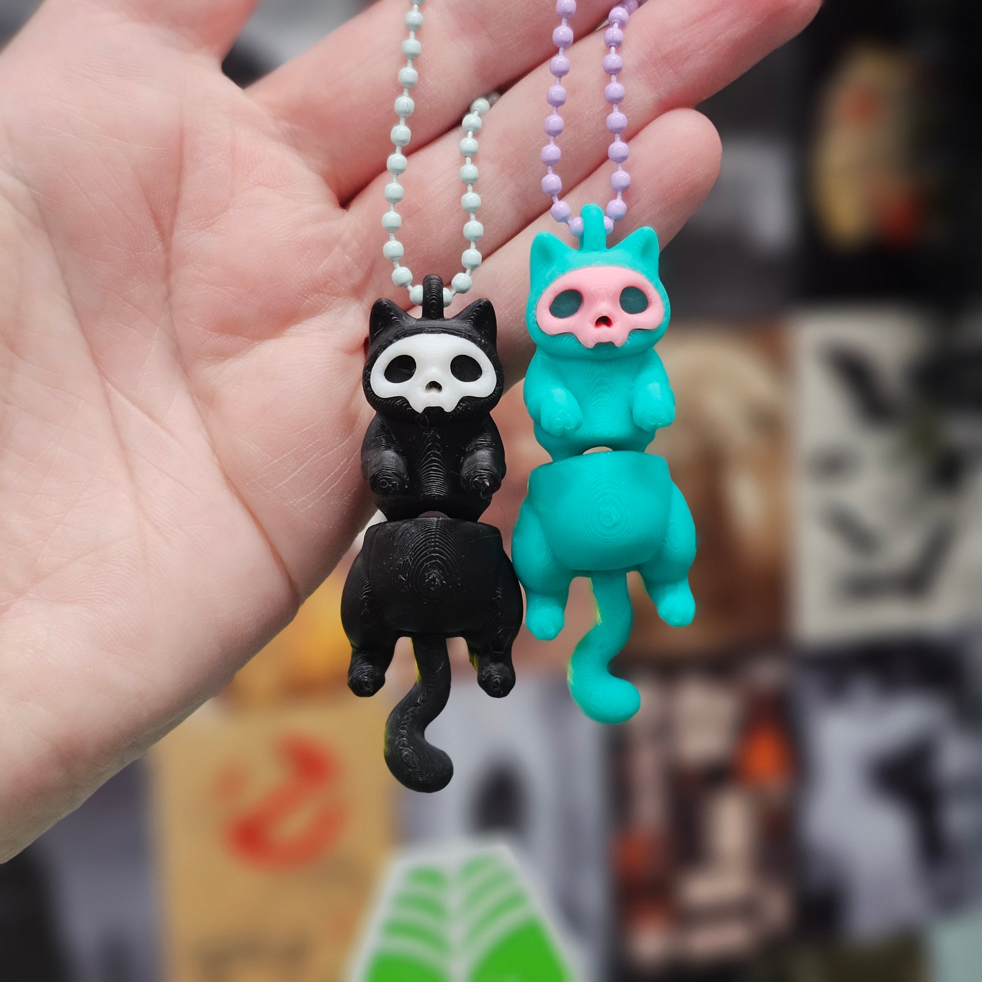 Two colorful keychains, one black and white cat-like design and the other turquoise with pink accents, held in a hand against a blurred background.