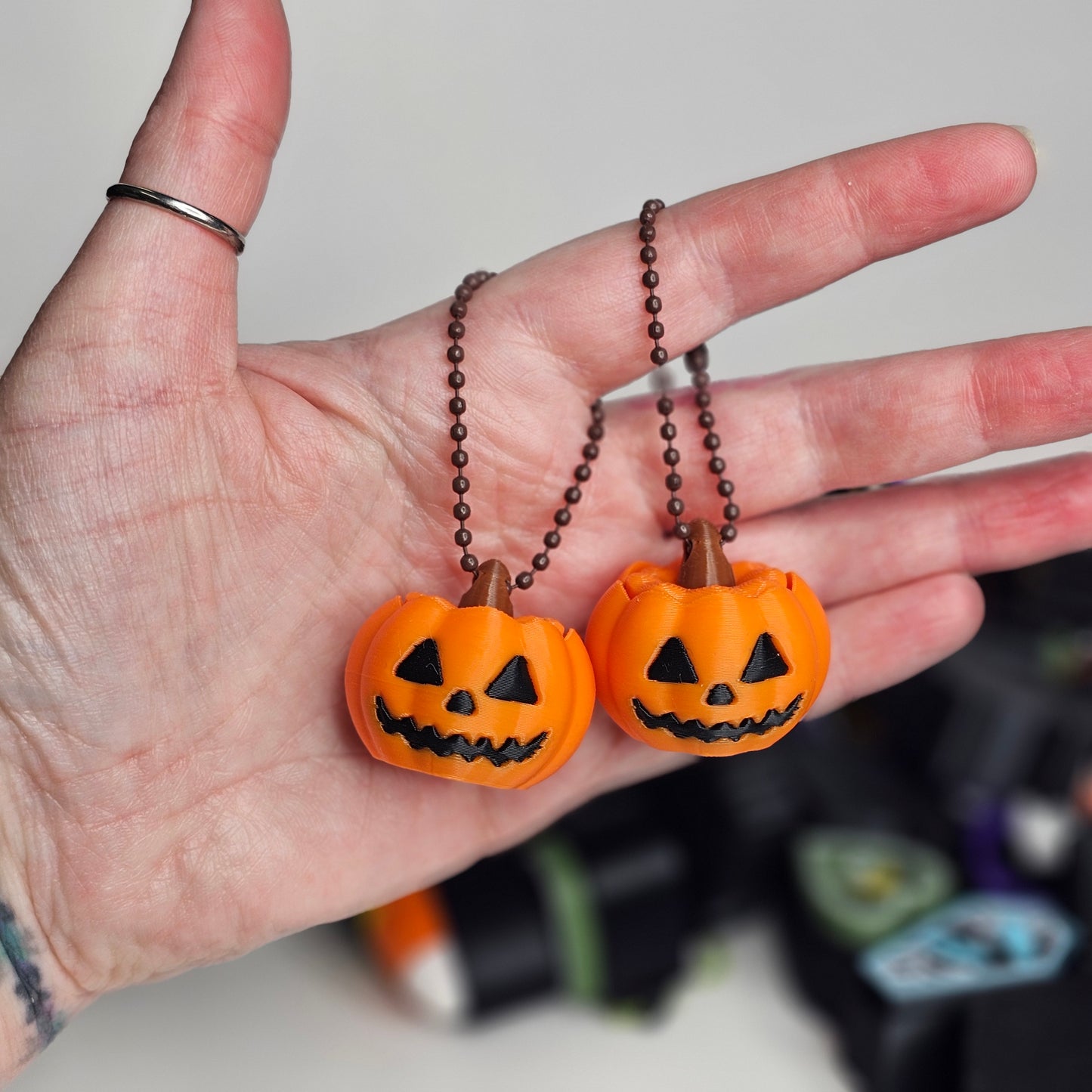 Two orange pumpkin-shaped keychains on a chain held in a hand with a blurred Halloween-themed background.